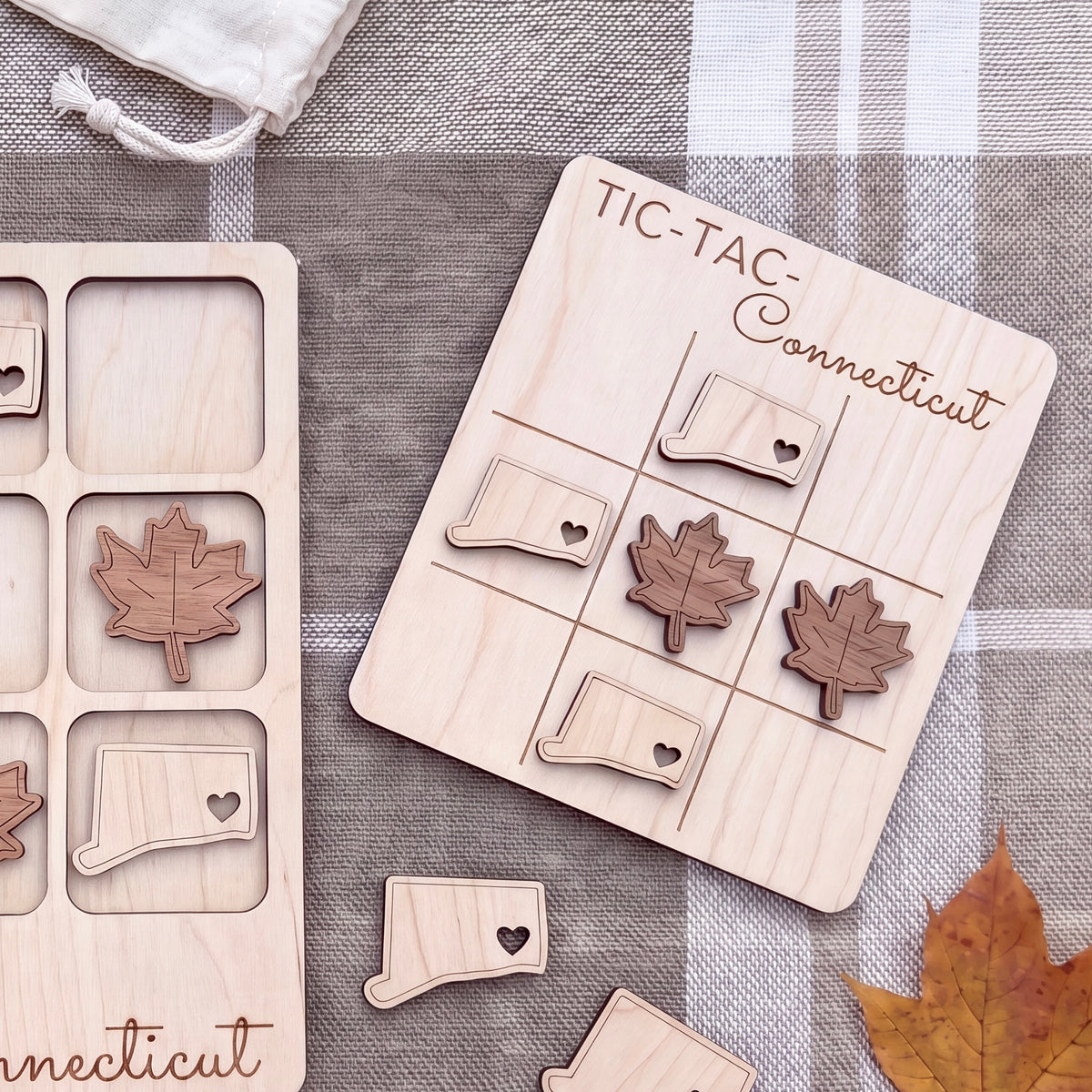 a wooden tic-tac game board with a Connecticut theme, including wooden leaves and state shapes, as well as a small wooden leaf on a gray cloth.