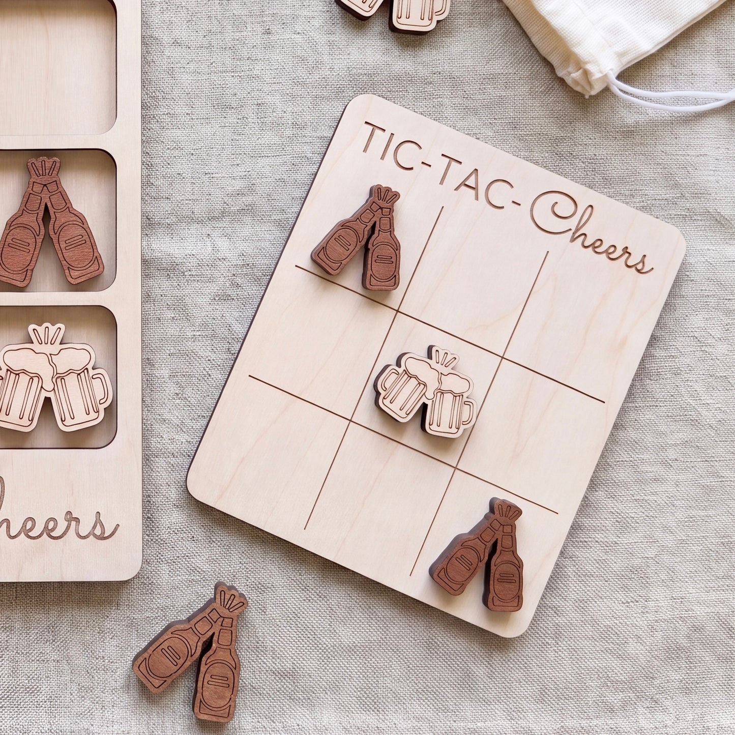 a wooden tic-tac game board with beer mug icons and a bag of wooden beer bottle icons on a beige cloth background.
