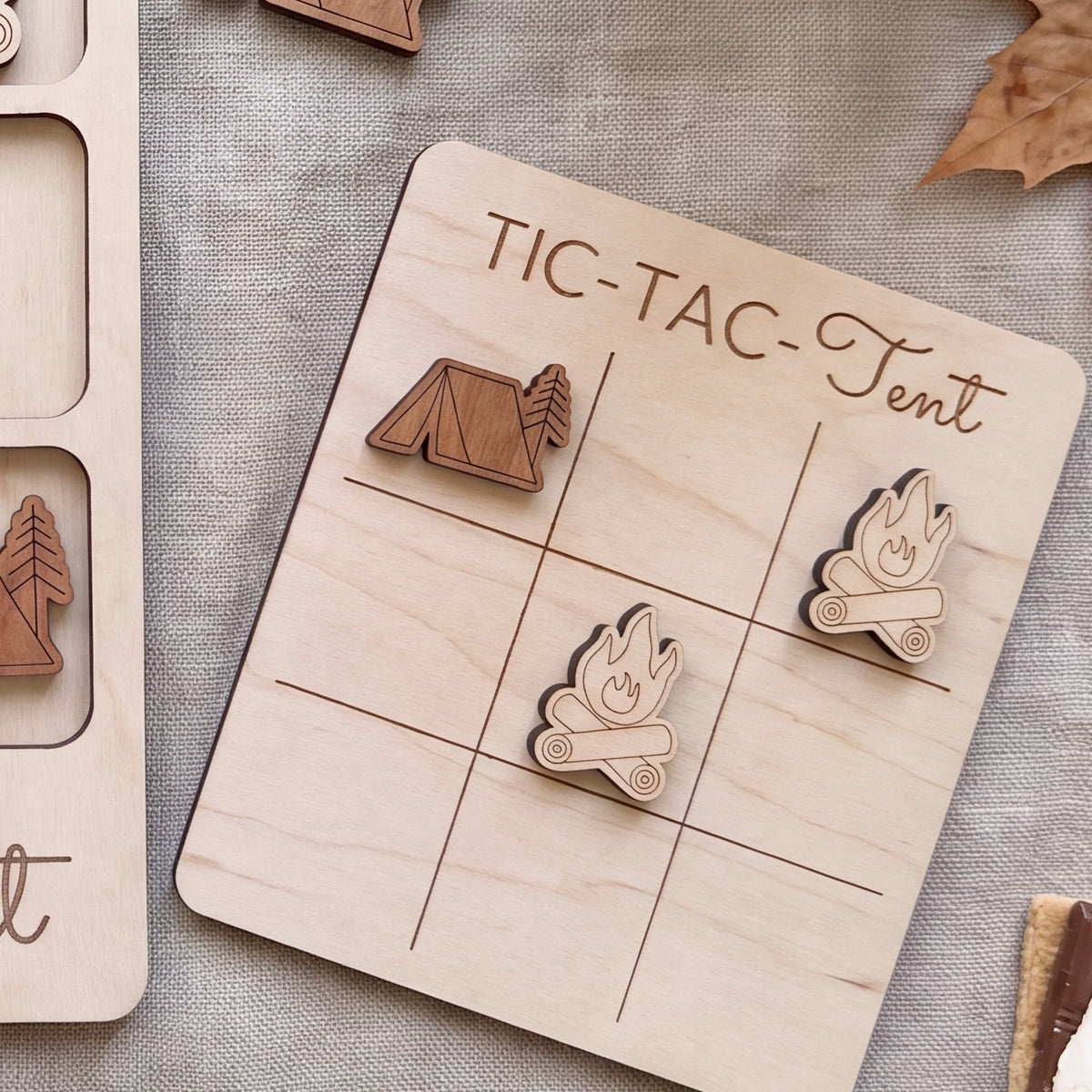 A wooden tic-tac game board with a tent and campfire motif, along with some wooden game pieces, on a grey fabric background.