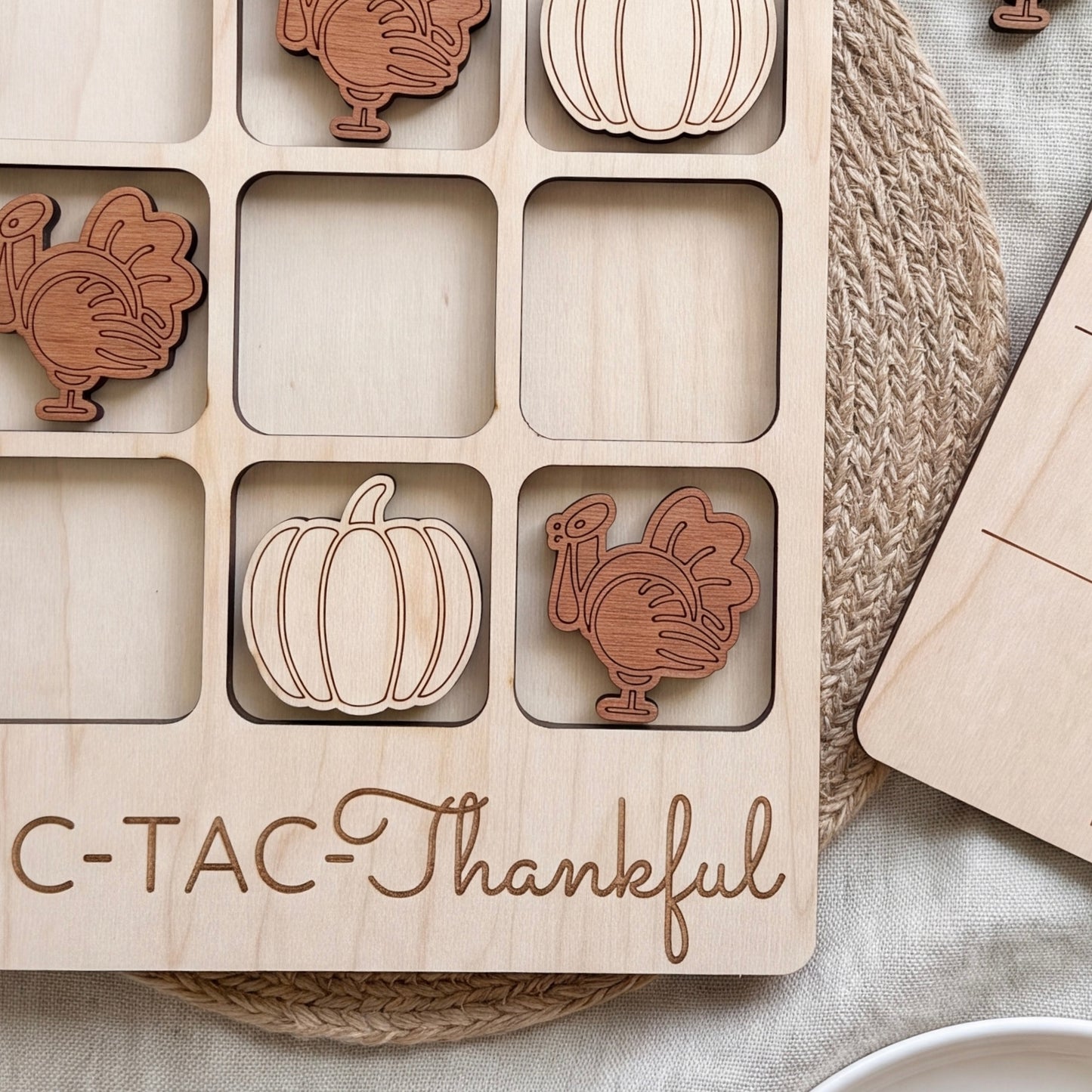 A wooden tic-tac-toe board with wooden turkey and pumpkin tiles, along with a wooden placemat with the phrase "C-TAC-Thankful" written on it.