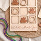 a wooden tic-tac game with wooden pieces shaped like beads and masks, placed on a woven placemat. There are also beads of different colors, including purple, green, and gold, scattered around the game.