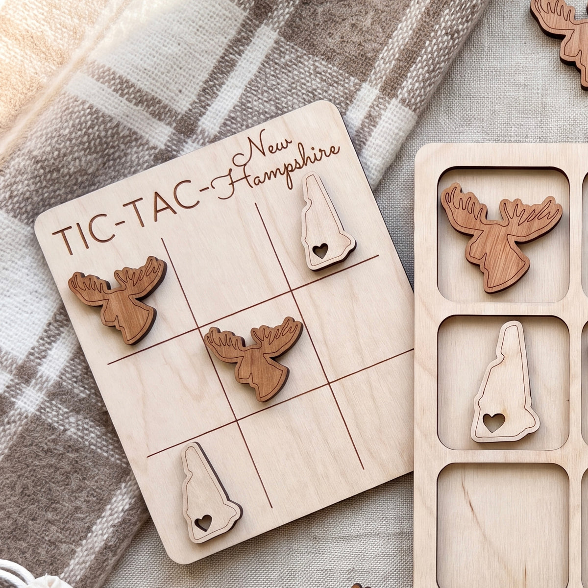A wooden tic-tac game board with wooden moose and heart-shaped pieces, placed on a blanket with a plaid pattern.
