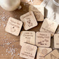 a collection of wooden coasters with various holiday-themed activities and items written on them, placed on a wooden surface with scattered white snowflakes.