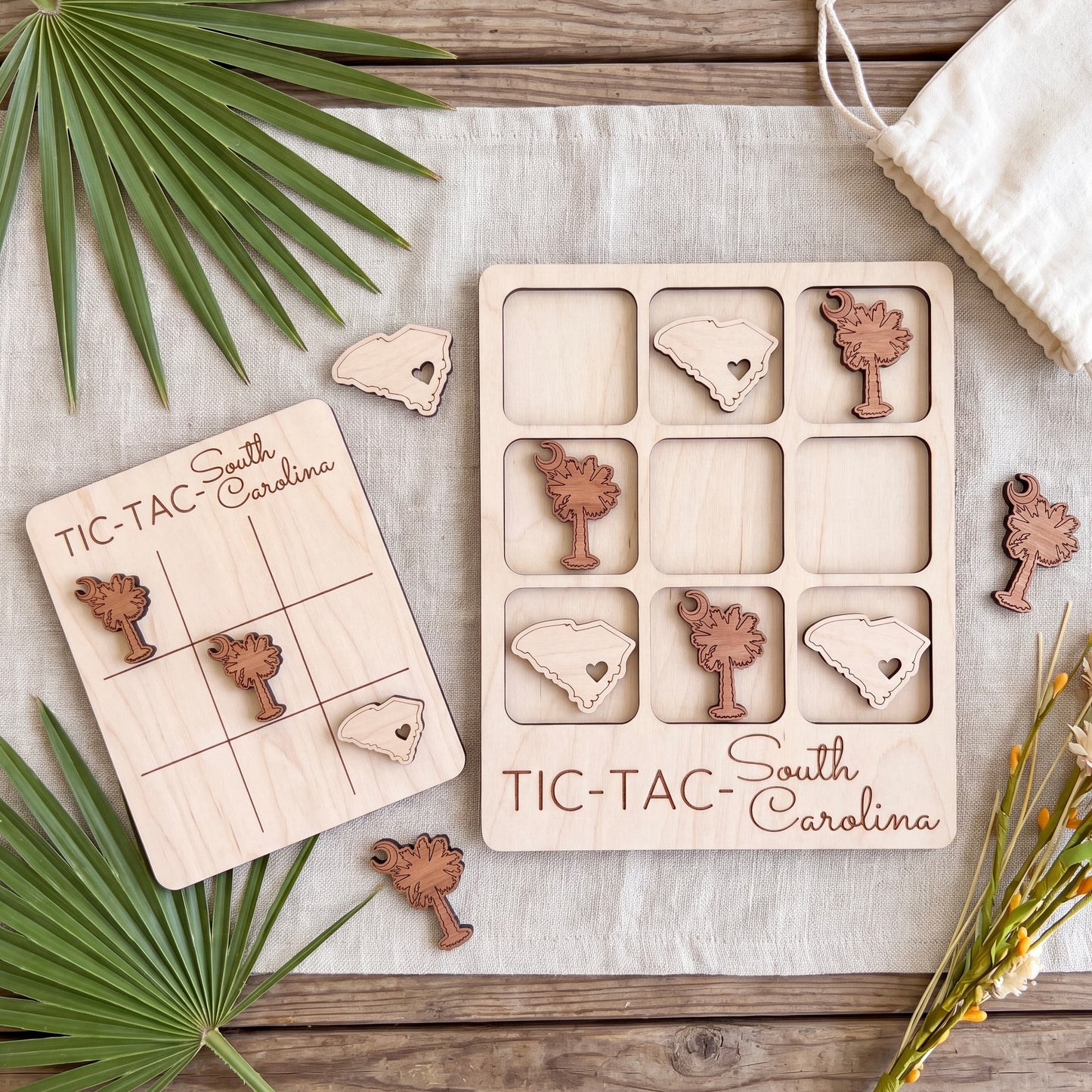 a wooden game board and wooden pieces shaped like the state of South Carolina, placed on a wooden surface with some green leaves and a white cloth.