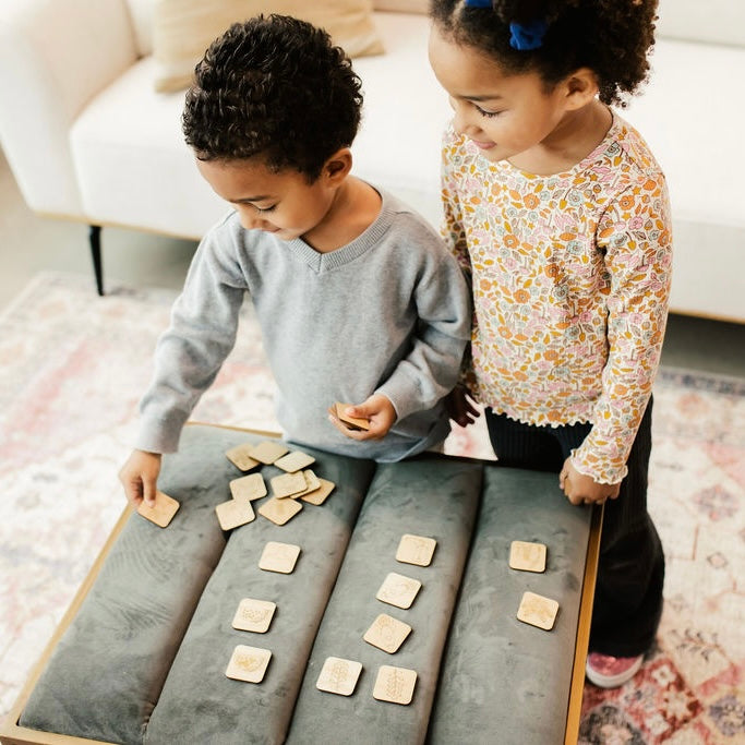 Two young children, a boy and a girl, are playing with wooden blocks on a gray couch.