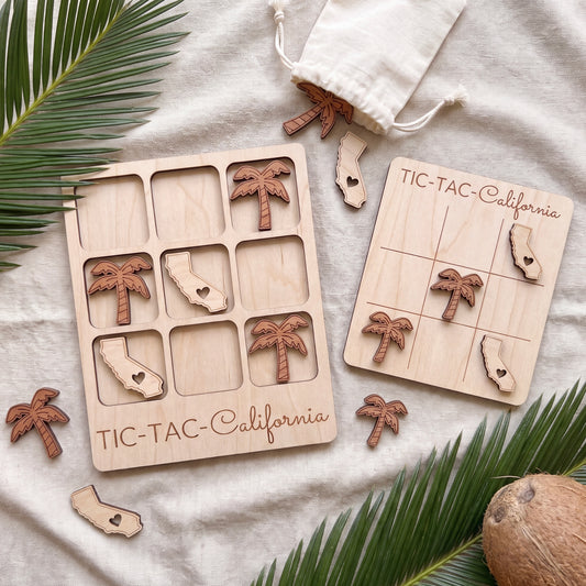 a wooden tic-tac game set with California tiles and a bag of wooden tiles, all placed on a white cloth with palm leaves and a coconut.