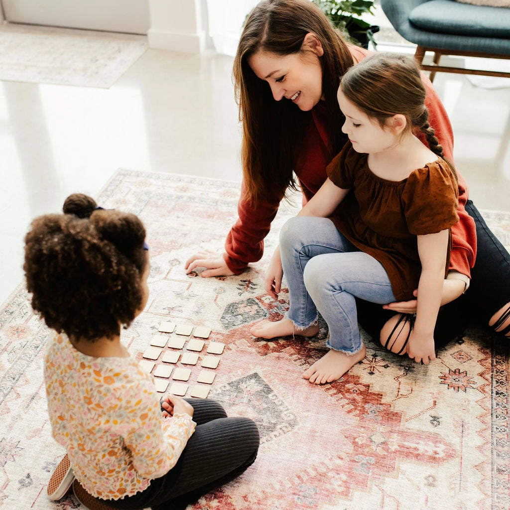 A woman and two young girls are sitting on the floor, playing a game with wooden blocks.