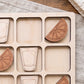 A wooden board with nine square compartments, each containing a wooden cutout of a glass and a slice of orange.