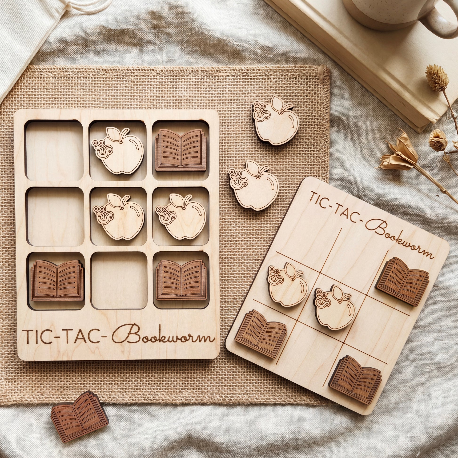 a wooden tic-tac board game called "TIC-TAC Bookworm" with wooden pieces shaped like books and apples. The game is placed on a beige cloth, and there are some additional wooden pieces and a cup visible in the background.