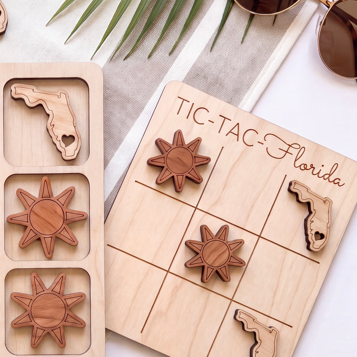 a wooden tic-tac game board with wooden pieces shaped like suns and a Florida state flag, along with a pair of sunglasses and a palm leaf.