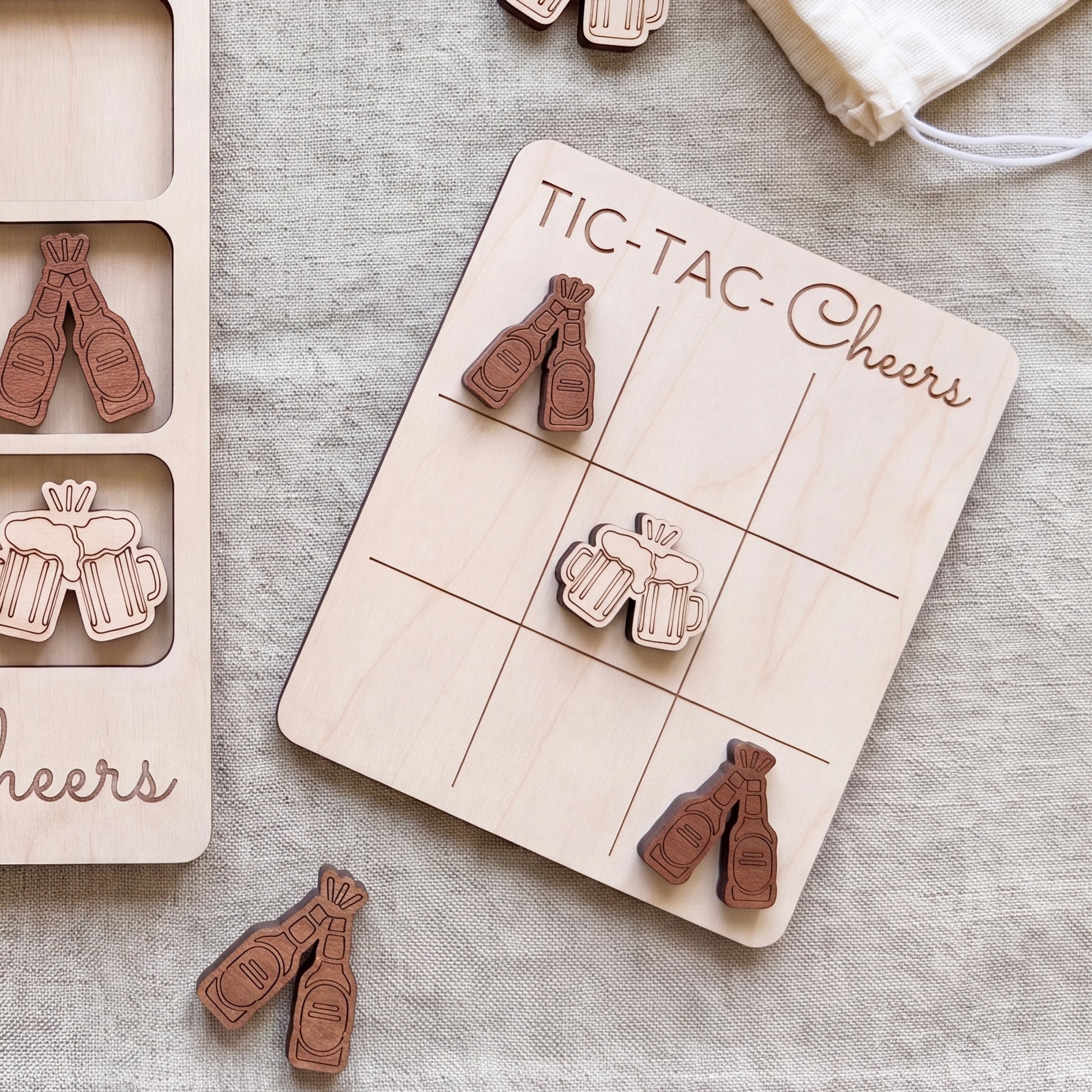 a wooden tic-tac game board with beer mug icons and a bag of wooden beer bottle icons on a beige cloth background.