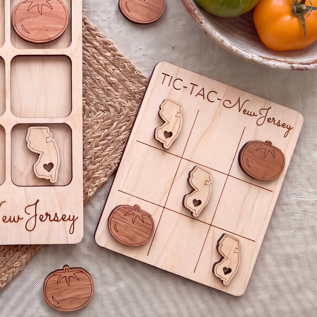 a wooden tic-tac game board with wooden pieces shaped like the state of New Jersey, as well as a wooden bowl containing tomatoes and a woven placemat.