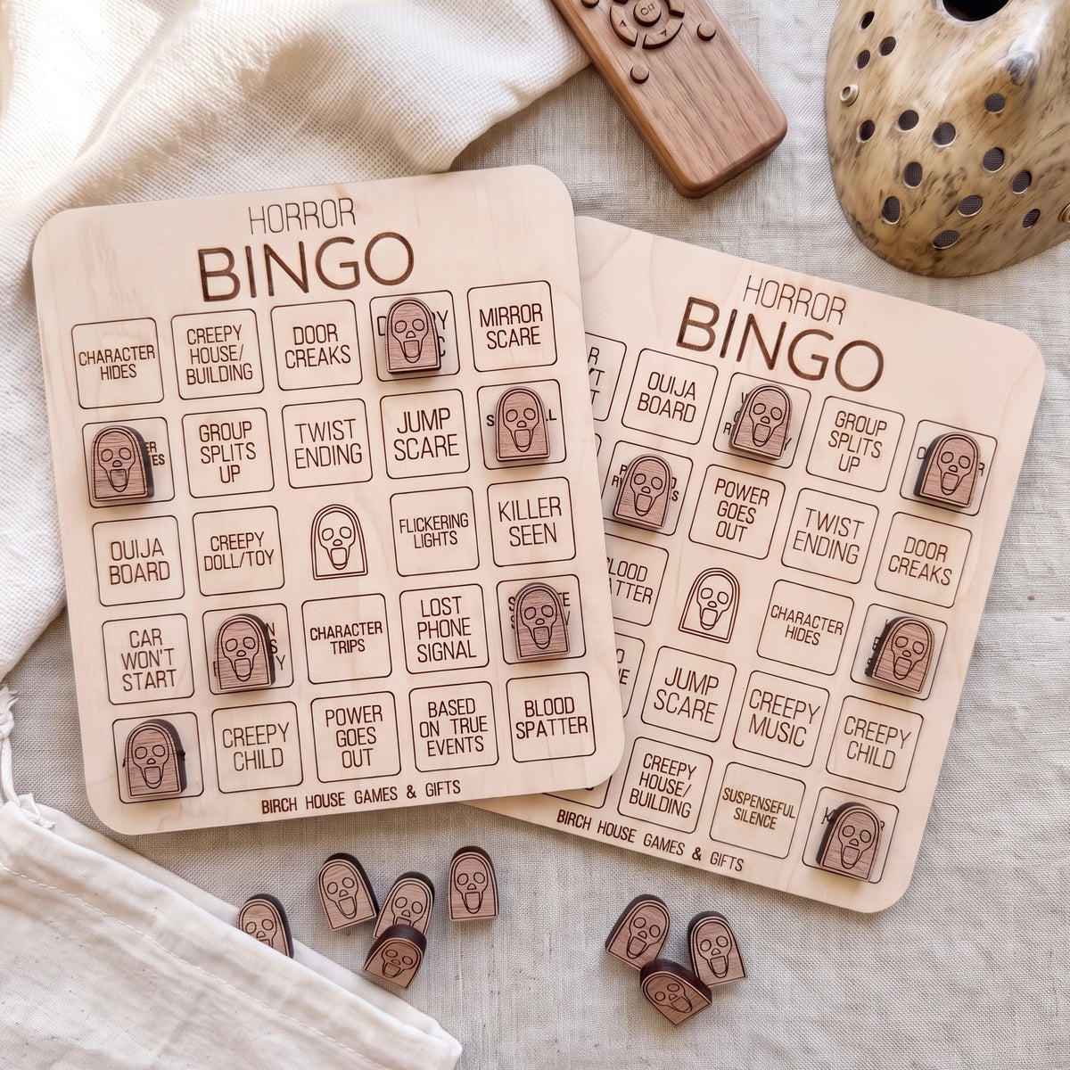 two wooden bingo cards with various scary-themed words and phrases printed on them, along with some wooden game pieces and a wooden remote control.