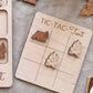 A wooden tic-tac game board with a tent and campfire motif, along with some wooden game pieces, on a grey fabric background.