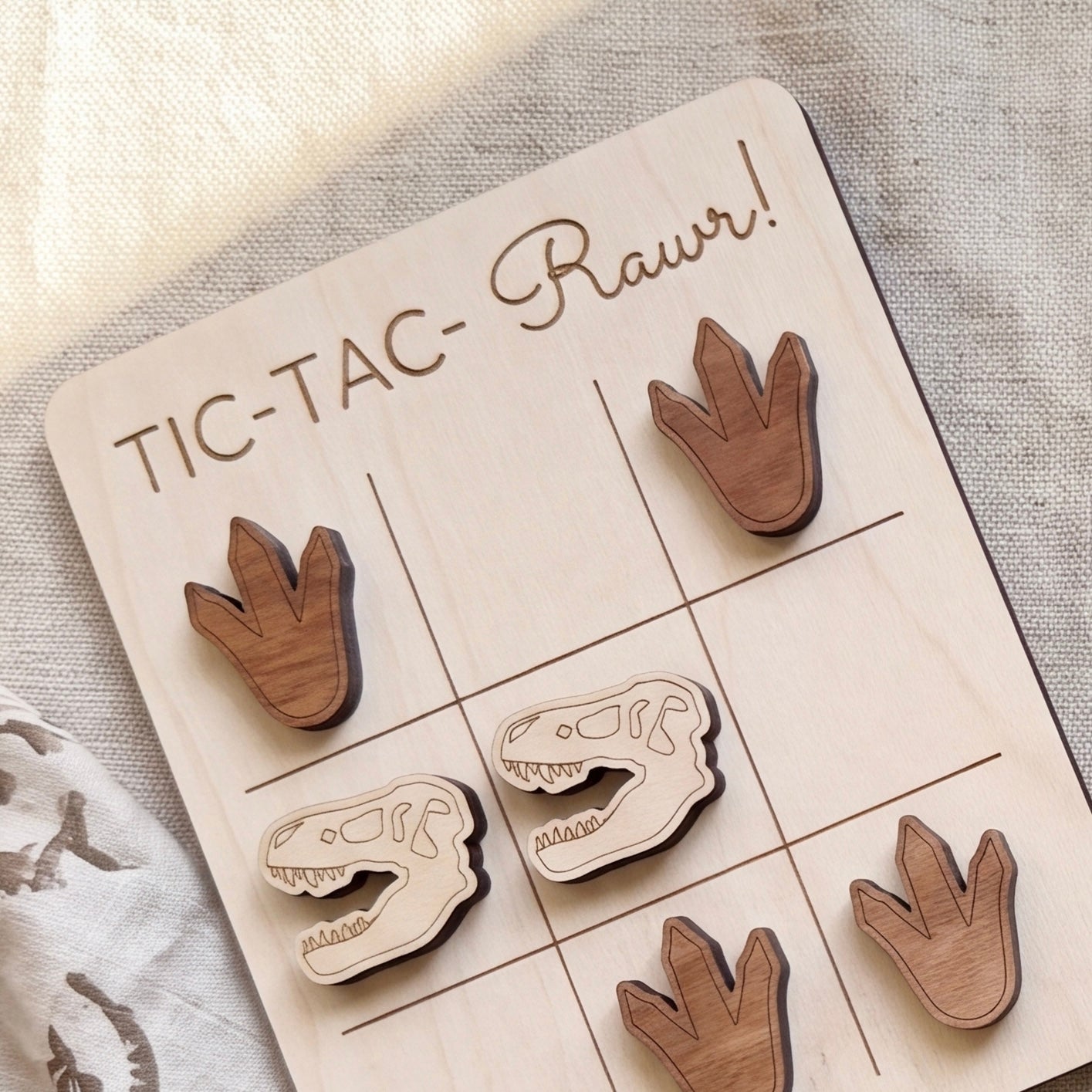 A wooden tic-tac game board with dinosaur claw and dinosaur skull tiles, along with the text "TIC-TAC-Rawr!" written on the board.
