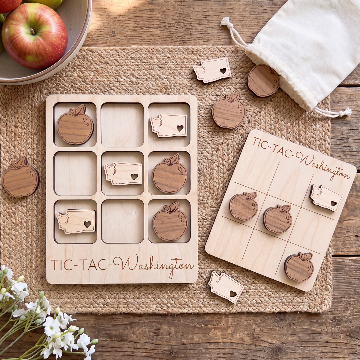 a wooden tic-tac game set with wooden pieces shaped like apples, placed on a wooden placemat. There is also a bowl of apples and a white cloth bag in the background.
