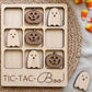 A wooden tic-tac-toe game board with carved pumpkin and ghost pieces, along with a spooky Halloween-themed game board and candy.