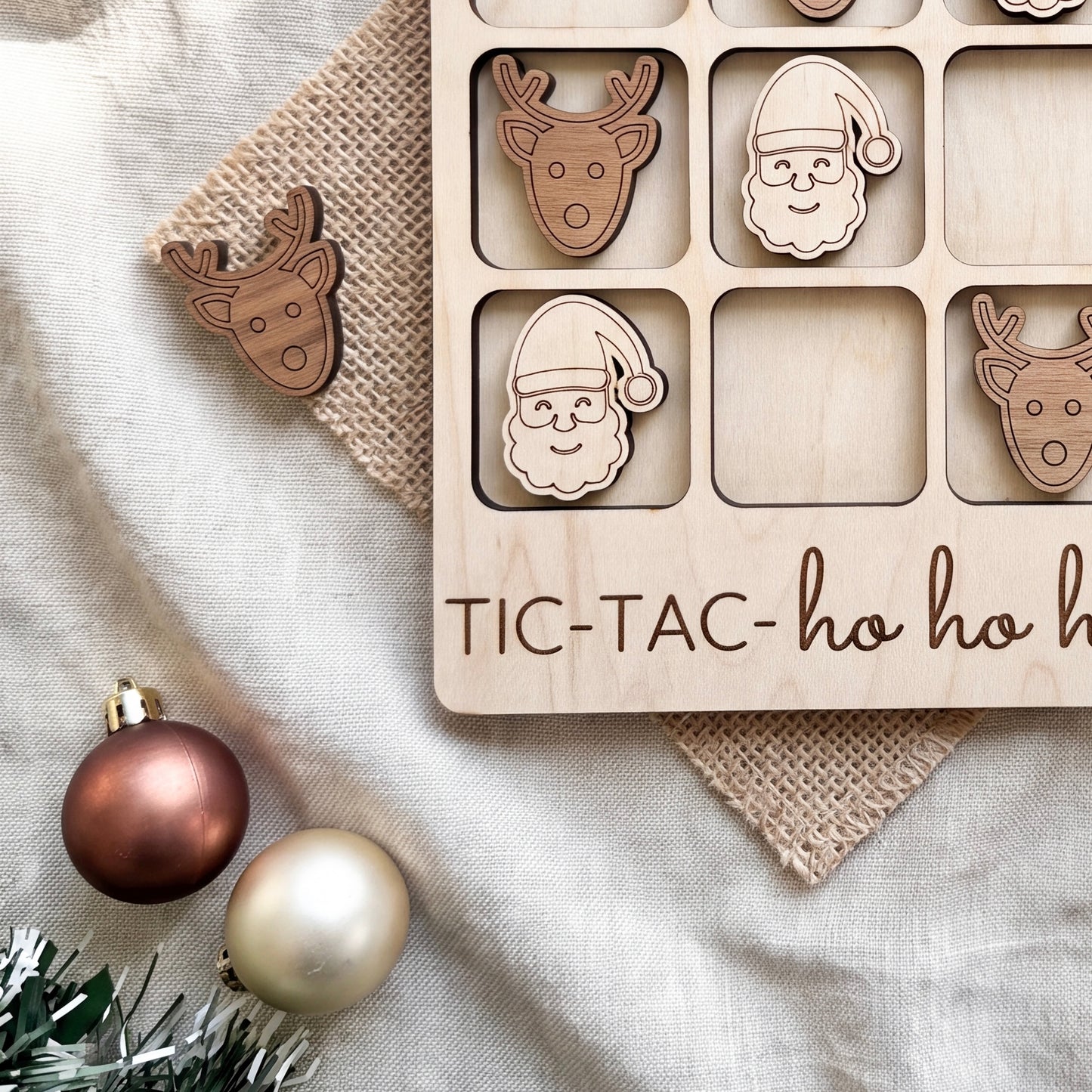a wooden tic-tac game with wooden pieces shaped like Santa Claus and reindeer, along with a Christmas ornament and a burlap cloth.