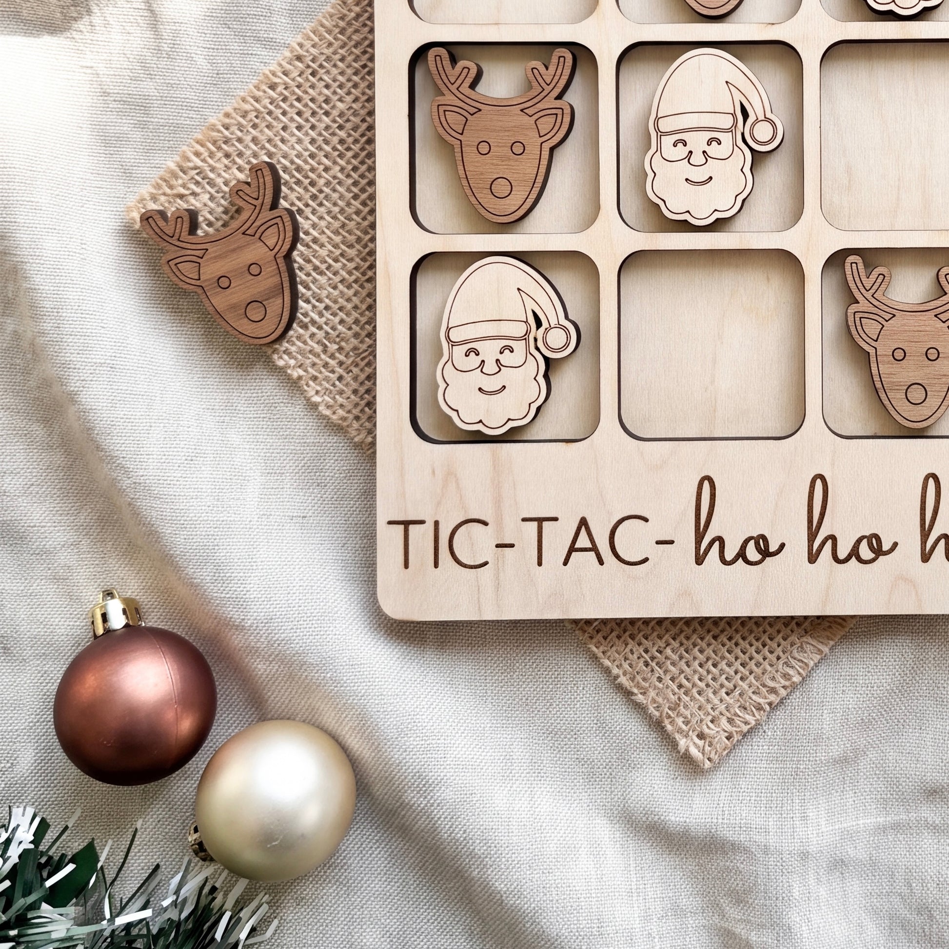 a wooden tic-tac game with wooden pieces shaped like Santa Claus and reindeer, along with a Christmas ornament and a burlap cloth.