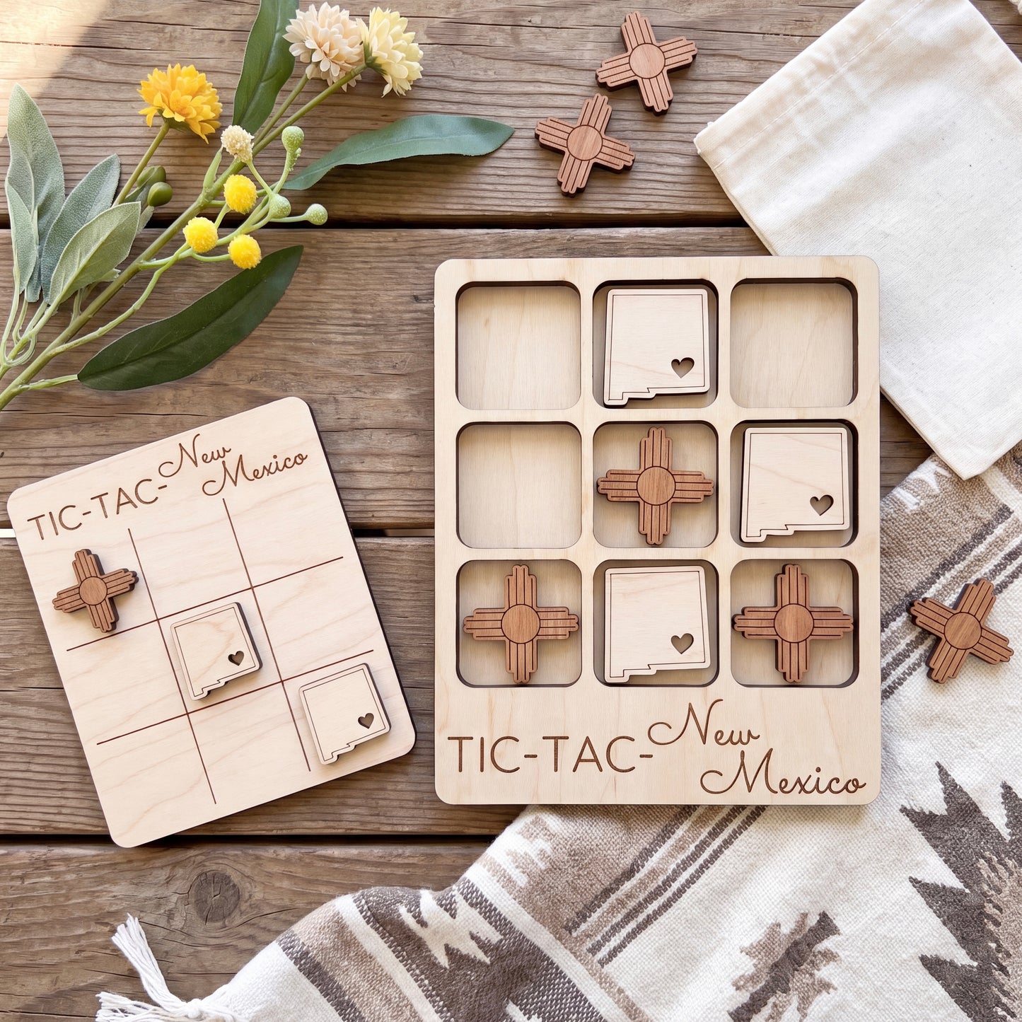 a wooden tic-tac game set with wooden pieces shaped like states in New Mexico, along with a wooden card that reads "TIC-TAC New Mexico". The game pieces are placed on a wooden surface, and there are some flowers and a white cloth in the background.