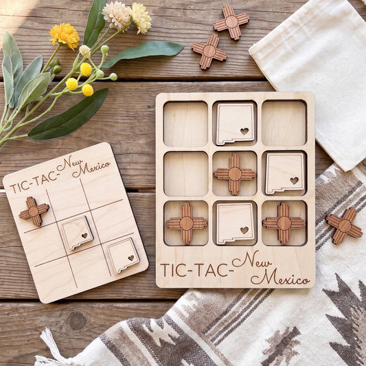 a wooden tic-tac game set with wooden pieces shaped like states in New Mexico, along with a wooden card that reads "TIC-TAC New Mexico". The game pieces are placed on a wooden surface, and there are some flowers and a white cloth in the background.