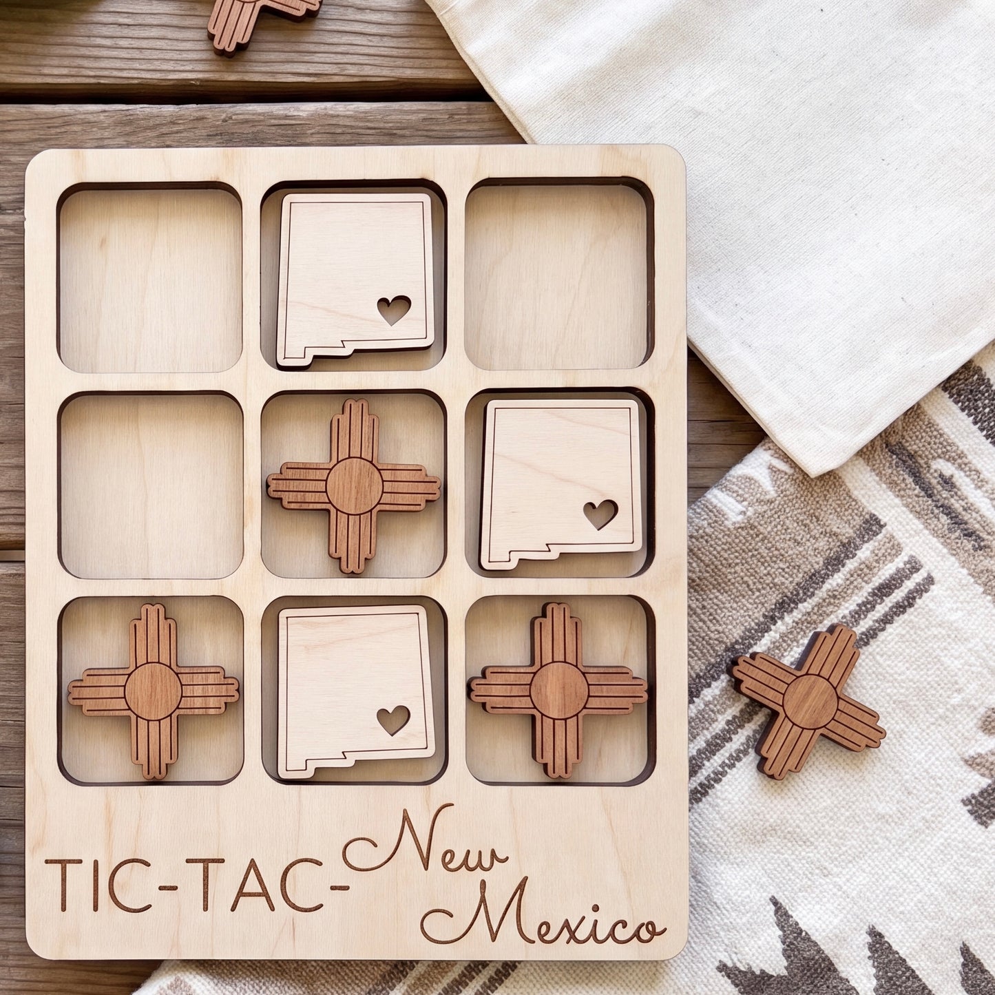 A wooden tic-tac game board with wooden pieces shaped like state flags, labeled "New Mexico", placed on a wooden surface.
