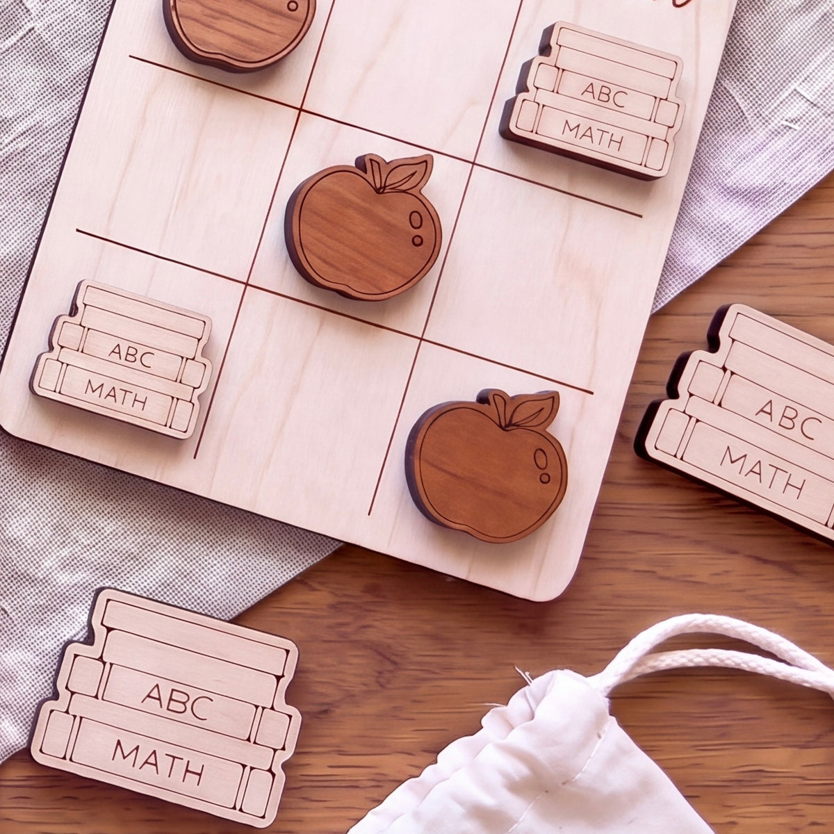 a wooden tic-tac-toe board with wooden apple and book shapes, along with a white cloth and a white bag.