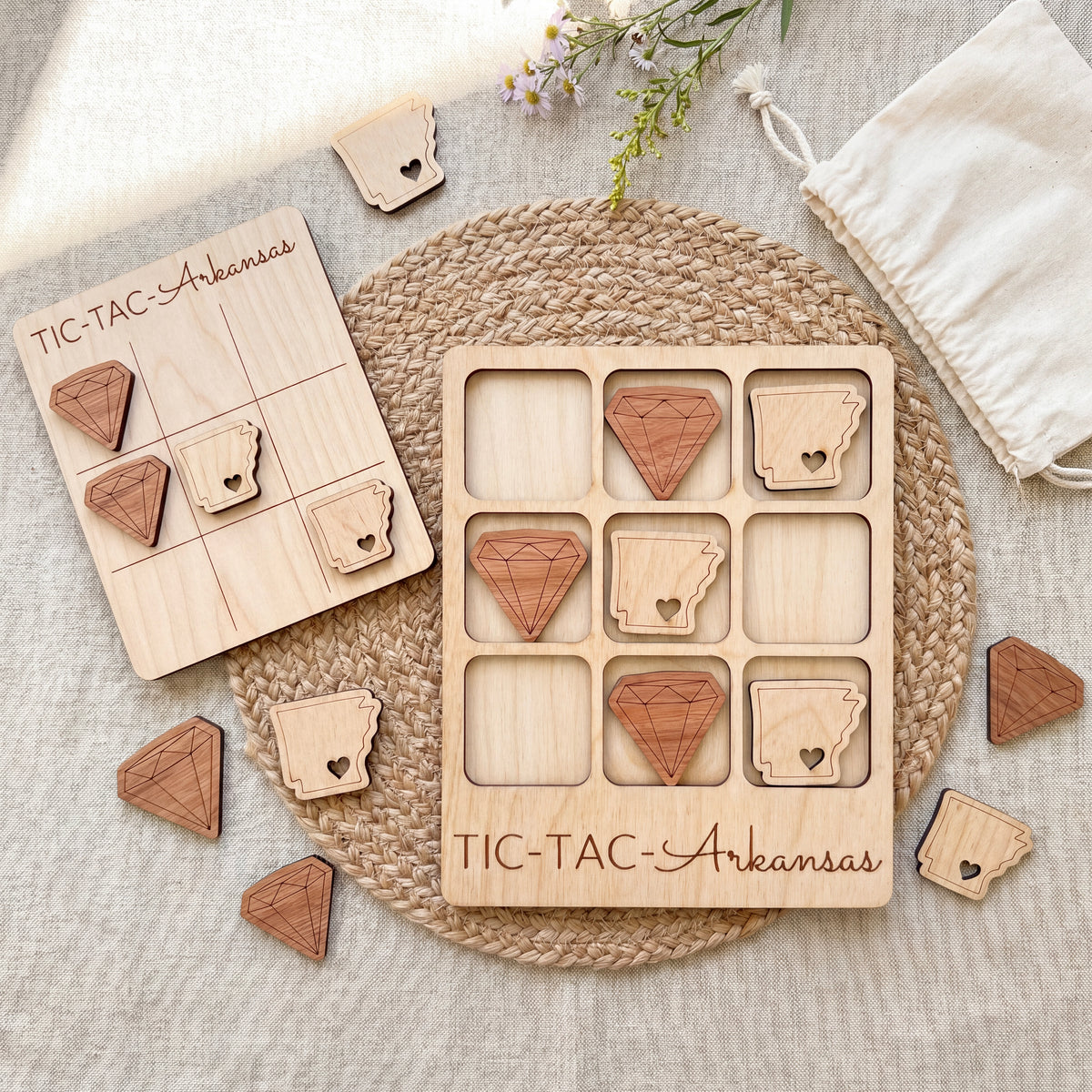 a wooden tic-tac game with wooden pieces shaped like diamonds and hearts, labeled "TIC-TAC-Aransas". The game is placed on a woven placemat, and there are additional wooden pieces scattered around the game.