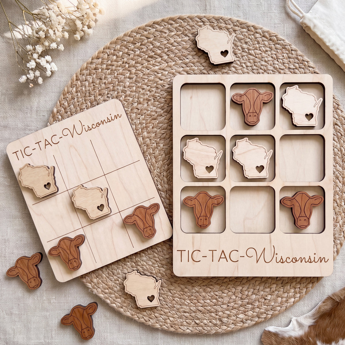 a wooden game board labeled "TIC-TAC-WISCONSIN" and a set of wooden cow-shaped tiles, all arranged on a woven placemat.