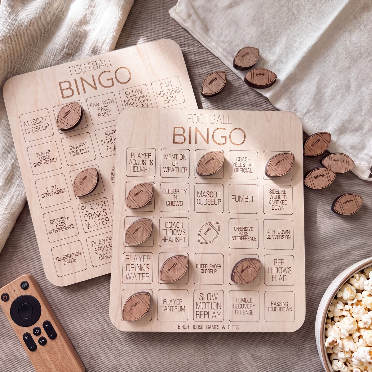 a wooden bingo board with football-related words and phrases, along with several wooden football-shaped game pieces scattered around. There is also a bowl of popcorn and a remote control on the table.