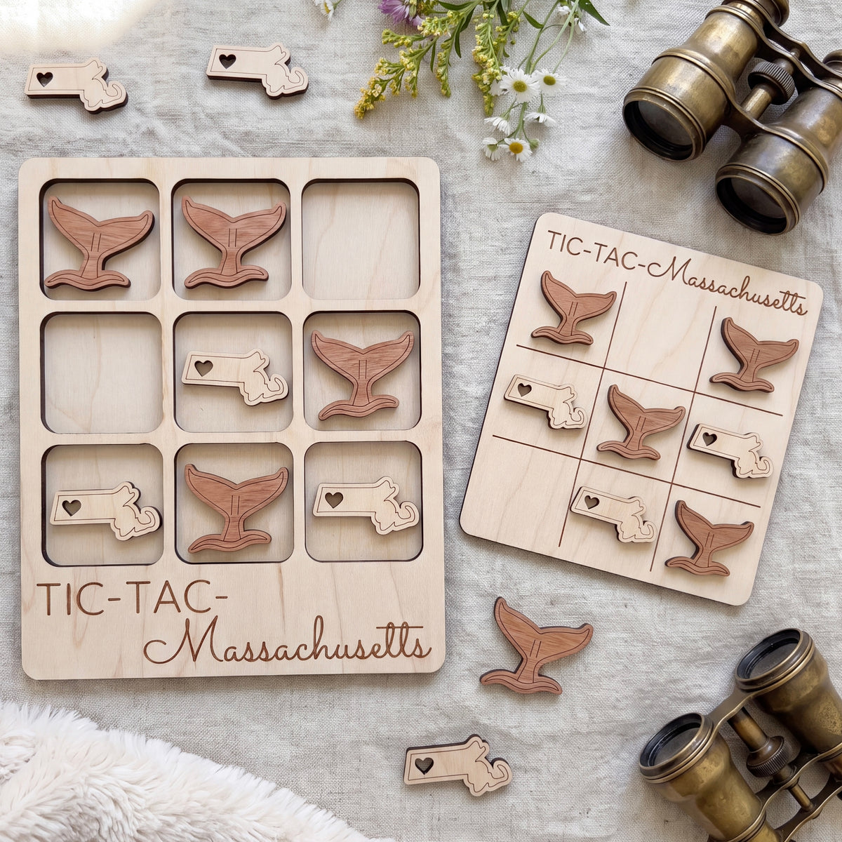 a wooden Tic-Tac game set with a Massachusetts theme, including a game board and pieces shaped like whale tails. There are also some binoculars and a floral arrangement in the background.