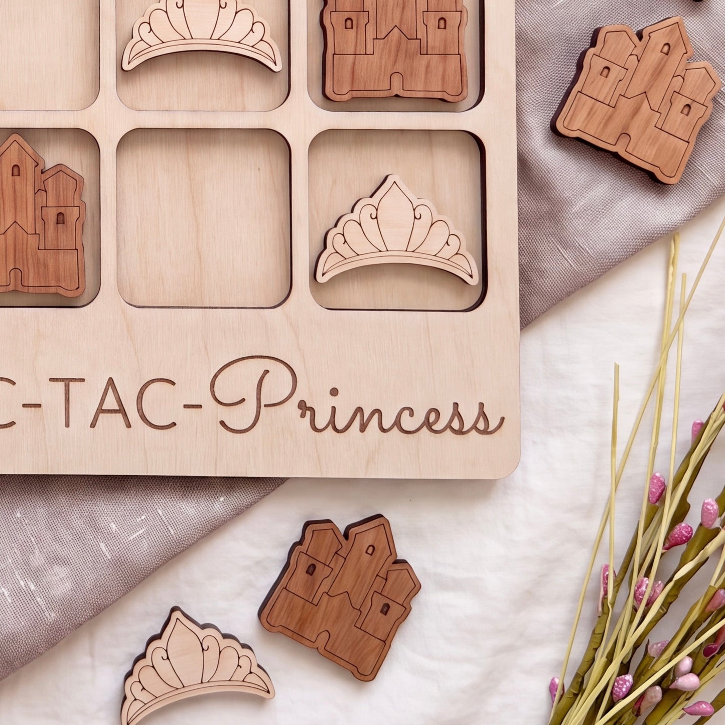a wooden tic-tac game with wooden crowns on it, along with some pink flowers and a gray cloth.