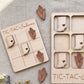 a wooden tic-tac game with a light wood texture and a light wood border. The game board is decorated with wooden cornucopia cutouts in the shape of the states of the United States, including Indiana. The game pieces are also made of wood and are placed on the game board.