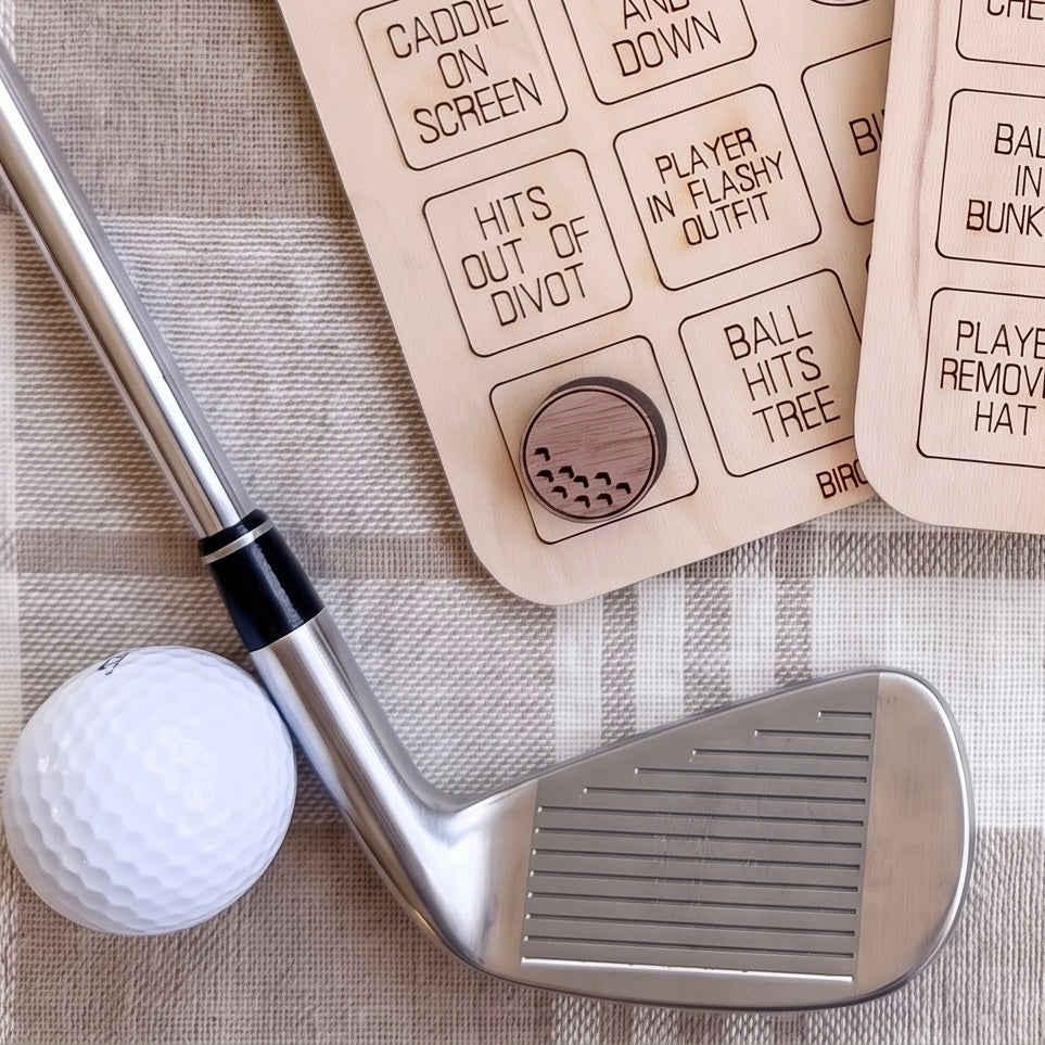 A golf club, a golf ball, and a wooden board with various golf-related labels are arranged on a textured surface.
