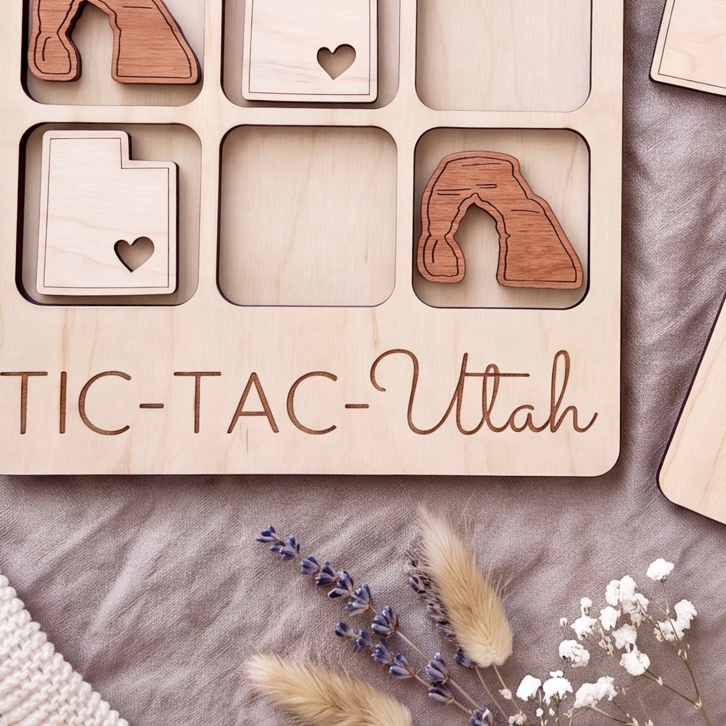 a wooden tic-tac game with the word "Utah" written on it, along with some wooden shapes resembling states and a heart. The game appears to be made of wood and is placed on a surface with some flowers and a knitted item.