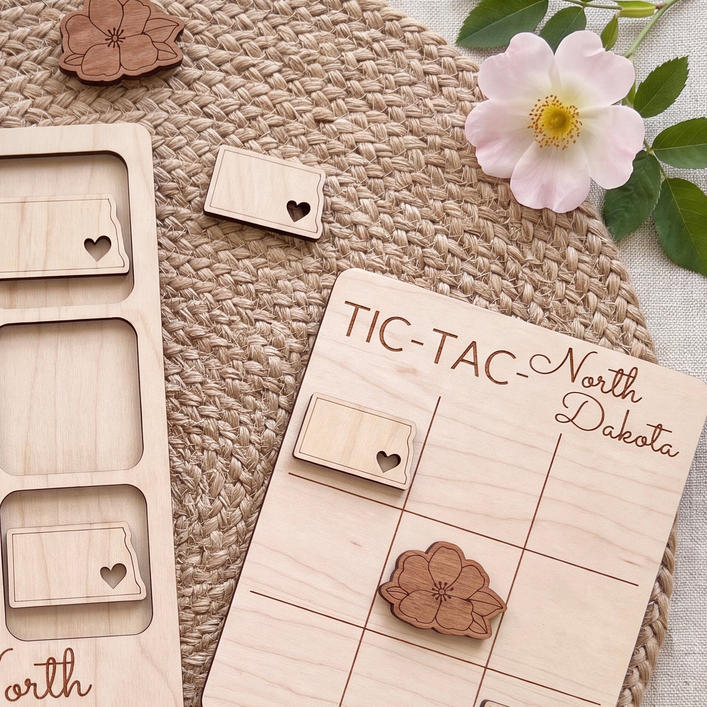 a wooden tic-tac game board with wooden pieces shaped like states, including those of North Dakota, placed on a woven placemat. There are also some wooden pieces with heart shapes and a pink flower, as well as a small wooden piece with a heart shape, all arranged on the placemat.
