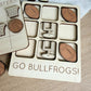 A wooden game board with various sports-themed cutouts, including a basketball, football, and a tic-tac-toe grid. The game board has a message that says "GO BULLFROGS!"