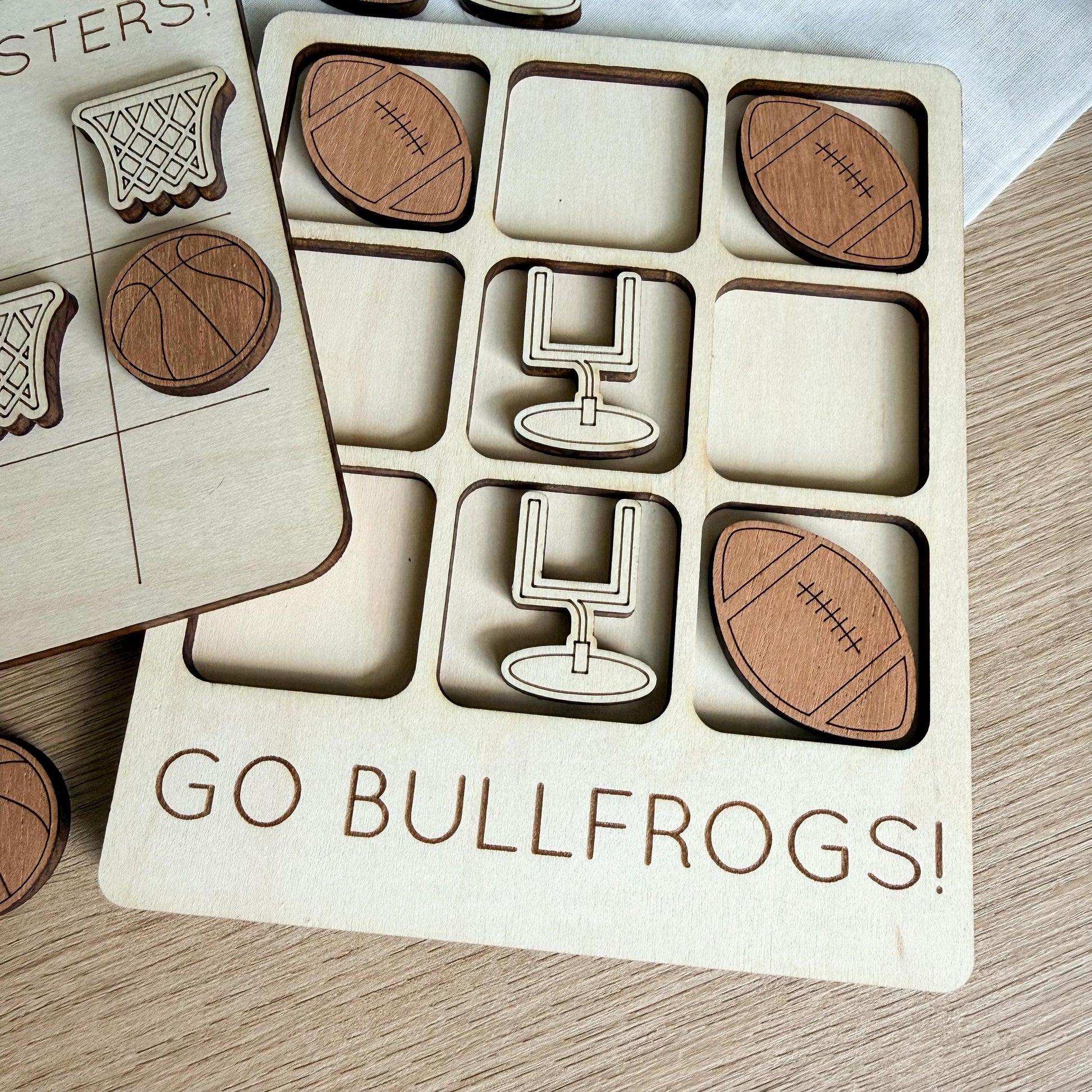 A wooden game board with various sports-themed cutouts, including a basketball, football, and a tic-tac-toe grid. The game board has a message that says "GO BULLFROGS!"