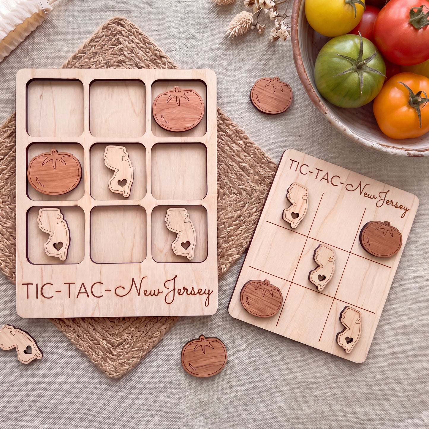 a wooden tic-tac game board and a wooden tic-tac card with wooden pieces, both labeled "TIC-TAC-New Jersey". The game board is placed on a woven placemat, and there is a bowl of tomatoes and some other fruits and vegetables in the background.