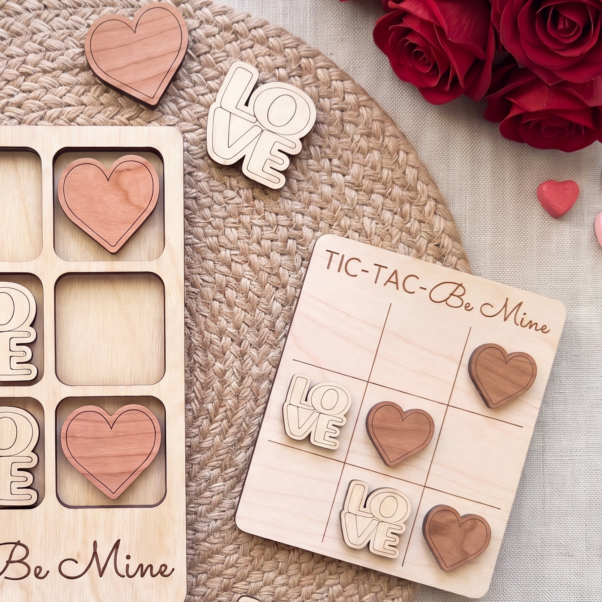 a wooden tic-tac game with the word "Love" written on it, along with several wooden heart-shaped tiles. There are also some wooden heart-shaped tiles and a wooden sign with the word "Love" written on it. The background includes a woven placemat and some red roses.