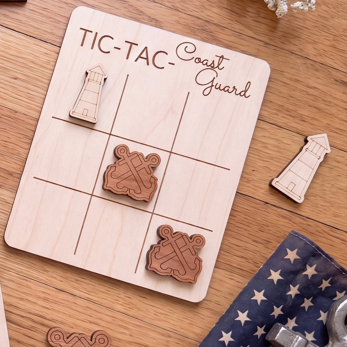 a wooden tic-tac game board with the words "TIC-TAC-Coast Guard" written on it, along with two wooden tic-tac pieces shaped like anchors. The game board is placed on a wooden surface, and there are some additional wooden objects, such as a star-patterned cloth and a small lighthouse-shaped object, in the background.