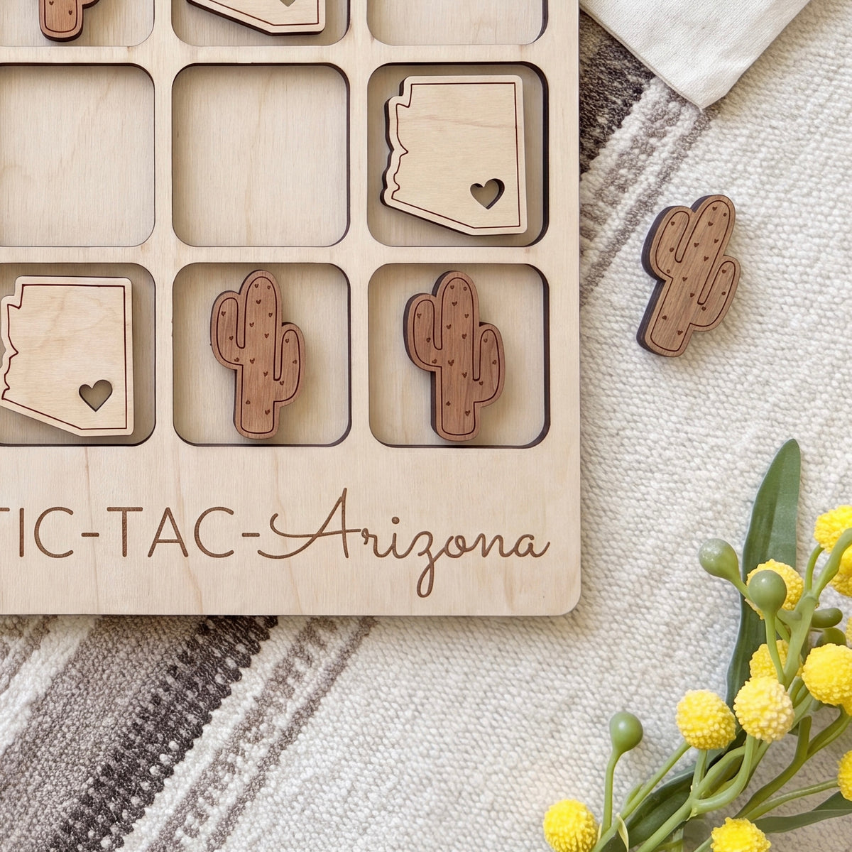 a wooden tic-tac game board with the word "TAC-TAC-ARIZONA" written on it, along with several wooden cactus or state-shaped tiles. There are also some yellow flowers or buds on the right side of the image.