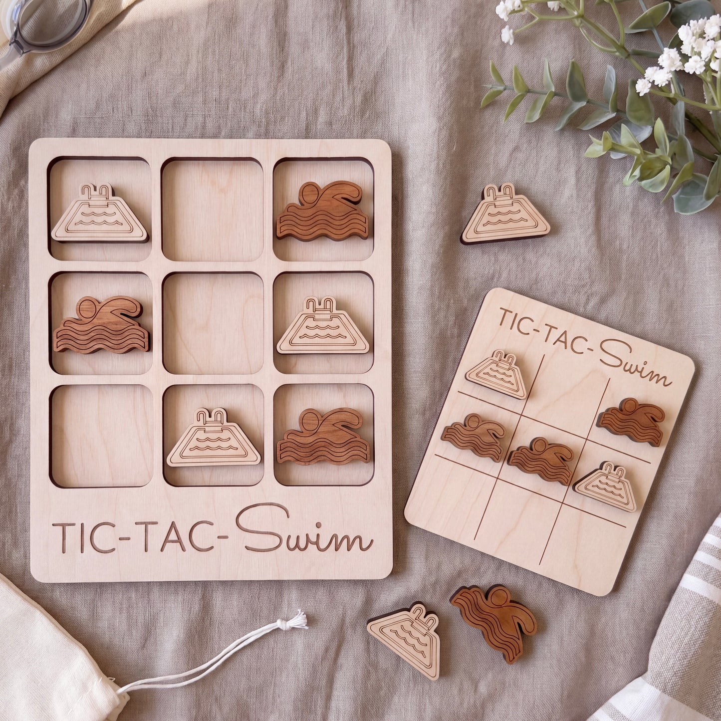 a wooden tic-tac-swim game with various swimming-themed tiles and a game board, along with some additional wooden tiles and a plant.