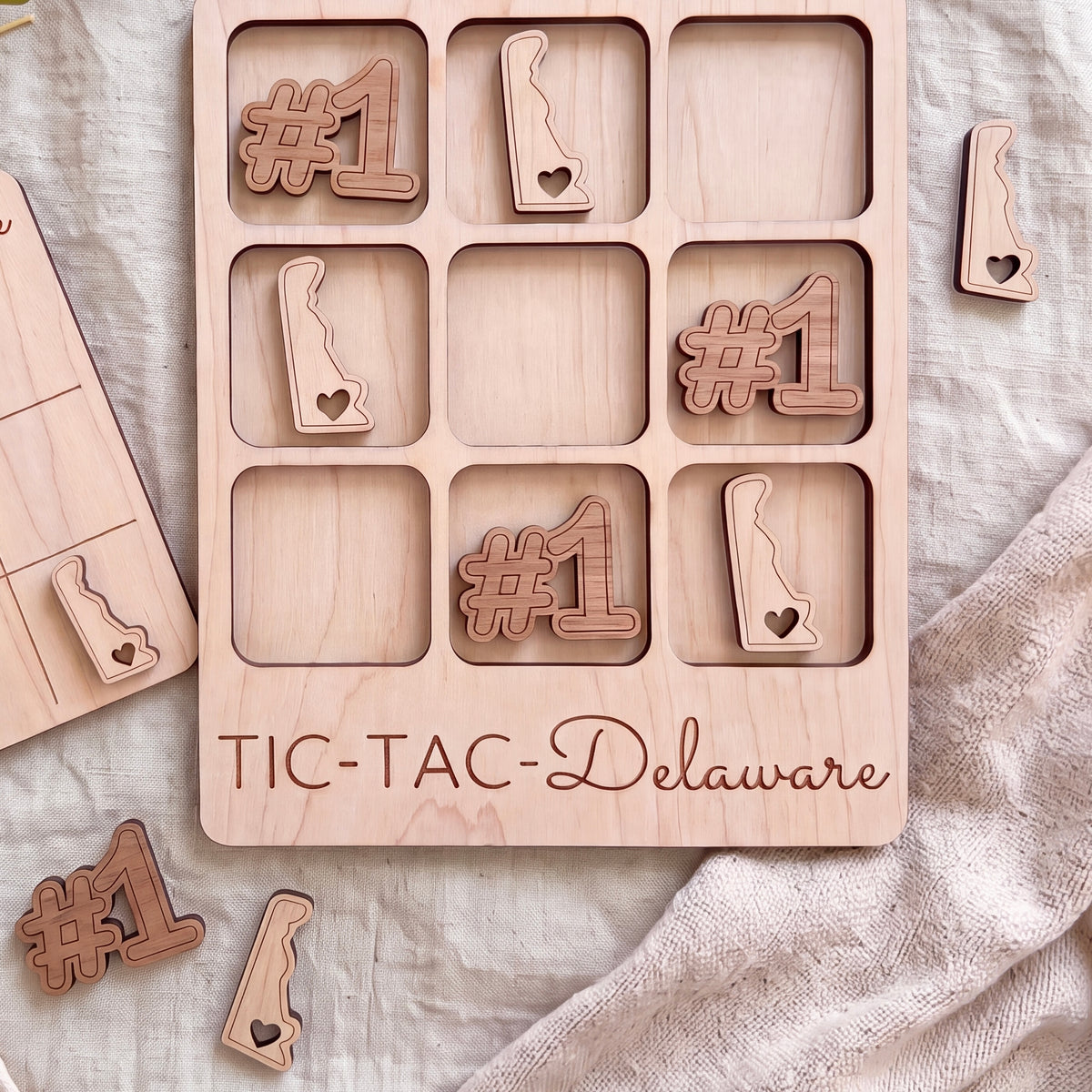 A wooden tic-tac game board with the word "TIC-TAC-Delaware" printed on it, along with several wooden pieces shaped like the state of Delaware.