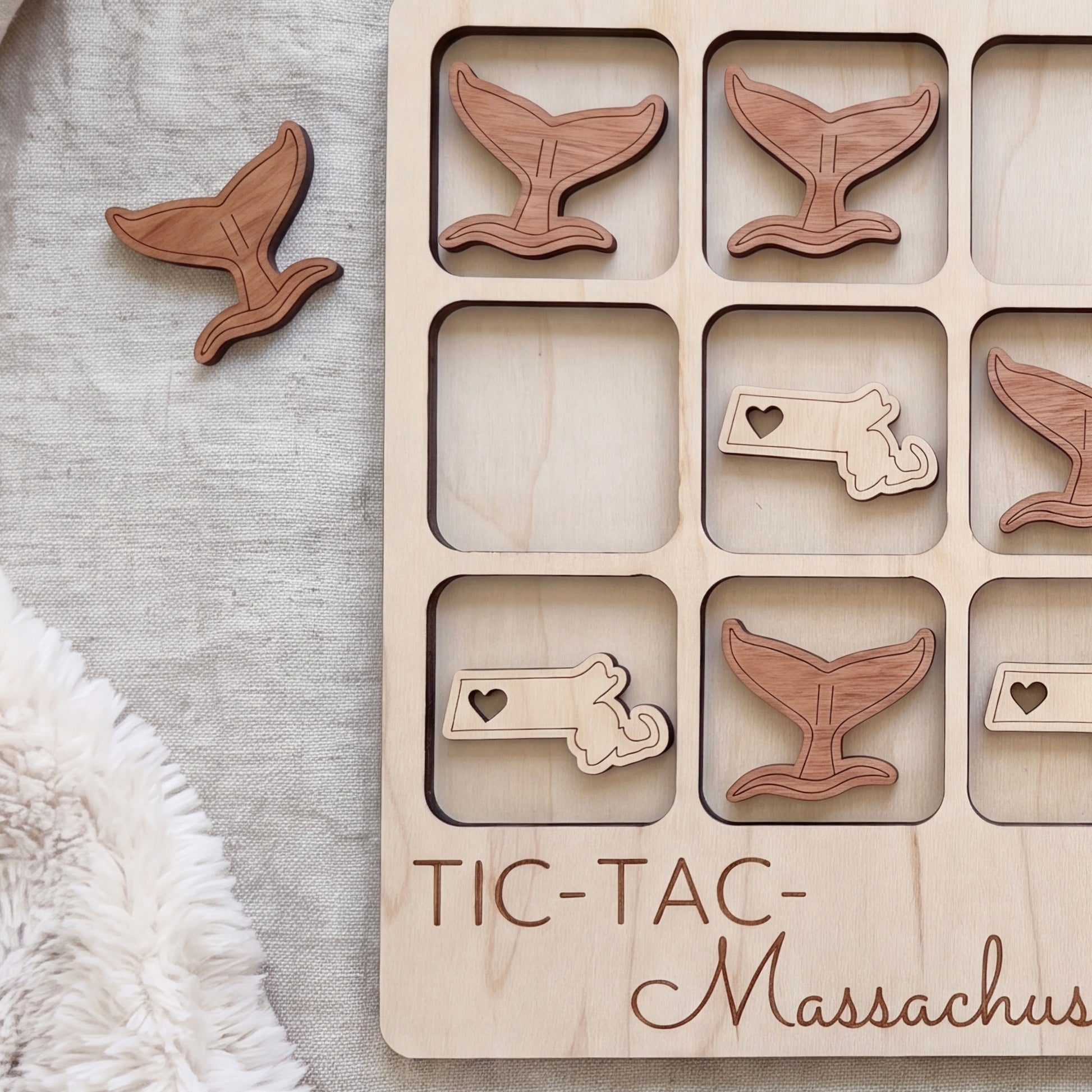 A wooden tic-tac game board with wooden tic-tac pieces shaped like whale tails, and the word "TIC-TAC-Massachusetts" written on the board.