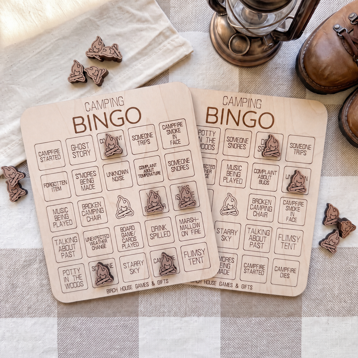 two wooden camping bingo cards with various camping-related words and phrases printed on them, along with some wooden bingo pieces and a pair of brown shoes.