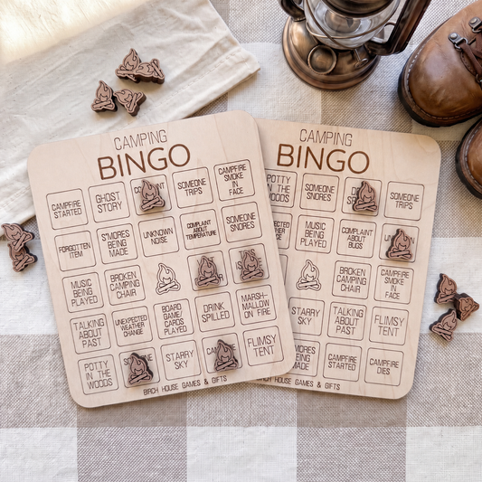 two wooden camping bingo cards with various camping-related words and phrases printed on them, along with some wooden bingo pieces and a pair of brown shoes.