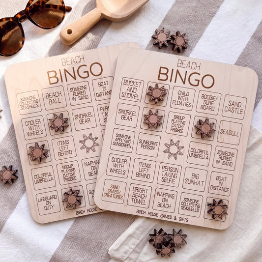 two wooden bingo cards with various beach-themed items and activities listed on them, along with some wooden bingo pieces and sunglasses on a striped towel.