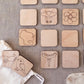 a collection of wooden coasters or game pieces with various simple designs, including a cow, flower, bird, and hot dog, arranged on a grey surface.