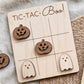 A wooden tic-tac game board with carved pumpkin and ghost pieces, along with the text "TIC-TAC-BOO!" written on the board.